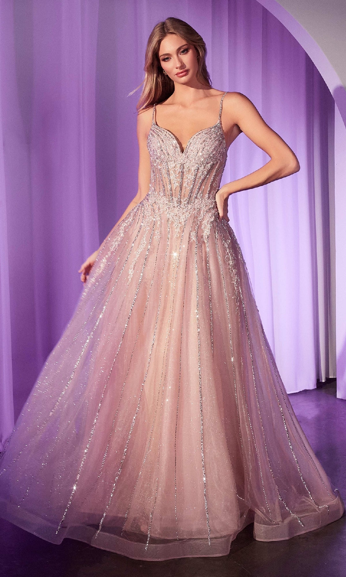 A woman stands indoors against purple draped curtains, wearing the Ladivine CD786 Crystal-Beaded Long Prom Dress—a sparkly light pink gown with thin straps and a fitted bodice.